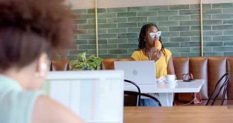 African american woman working remotely in cafe eating croissant while wearing sunglasses