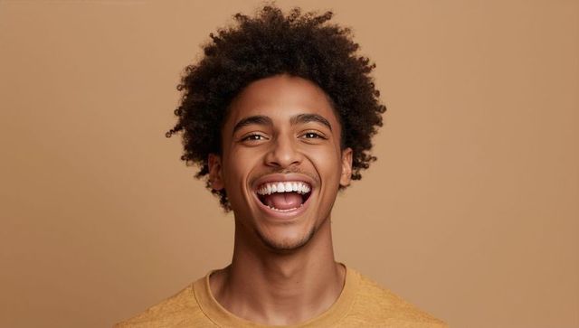 Smiling young man with curly hair laughing in mustard t-shirt closeup headshot on beige background