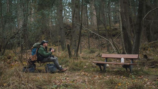 Solo hiker resting, contemplating with backpack and camera on bench in autumn forest glade