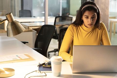 Focused indian professional woman working on laptop in modern open-plan office with coffee