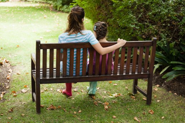 Rear view of woman and girl sitting on wooden bench at backyard