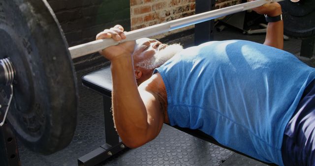 Senior man lifting a barbell while lying on a bench in a gym environment. Useful for illustrating senior fitness, weight training guides, health and wellness websites, and fitness program ads.