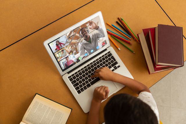 Young student participating in online learning using a laptop, with open books and colored pencils around. Ideal for educational websites, e-learning platforms, articles about remote education, and child development studies.