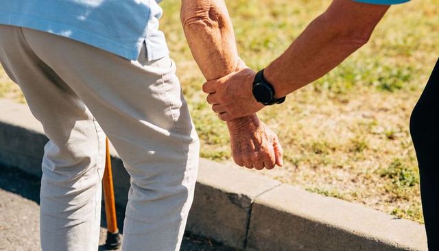 Caregiver helping elderly person up from curb holding forearm and cane in sunlit area