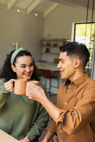 Young couple clinking matching mugs at sunlit kitchen table sharing cozy morning moment