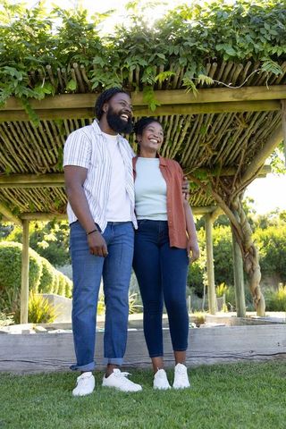 Smiling black couple standing under vine-covered pergola enjoying sunny garden moment together