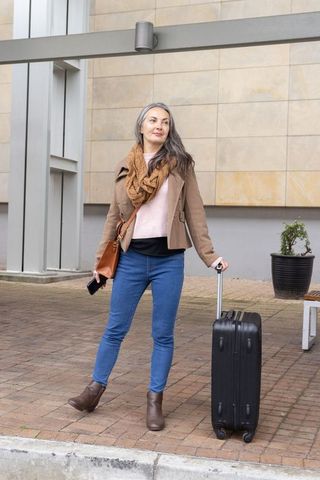 Mature woman checking phone while pulling rolling suitcase in urban plaza