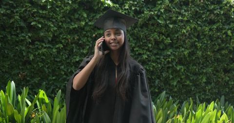 Graduating student talking on phone wearing cap and gown in lush green courtyard