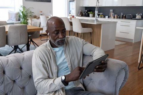 Senior african man relaxing on tufted sofa using tablet and earbuds in open-plan home