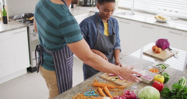 A young couple is preparing a healthy meal together in a modern kitchen. They are chopping fresh vegetables such as carrots, peppers, and cucumbers. They both wear aprons and focus on cooking tasks. This image is perfect for promoting healthy eating, lifestyle blogs, cooking classes, and kitchenware advertisements.