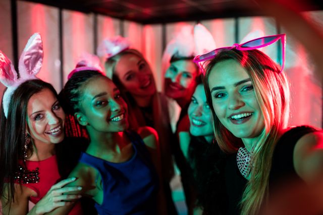 Group of friends enjoying a night out at a bar, posing for a selfie with smiles and bunny ears headbands. Neon lights create a vibrant atmosphere. Perfect for themes related to friendship, nightlife, celebrations, and social gatherings.