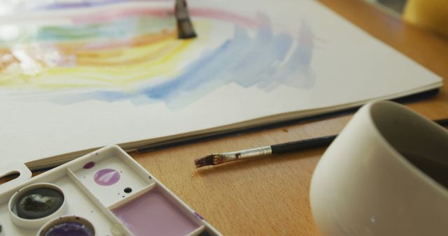 This image showcases a close-up of watercolor painting supplies including a paintbrush and a painter’s palette with various colors. The scene captures part of a watercolor artwork featuring vibrant and smooth transitions of colors on paper. Next to the supplies, there is a coffee cup on the wooden table. This high-quality image is perfect for art-related content, creative process visuals, or educational materials demonstrating the use of watercolors. It can also be used in blogs, websites, and advertisements focused on art, creativity, and hobbies.