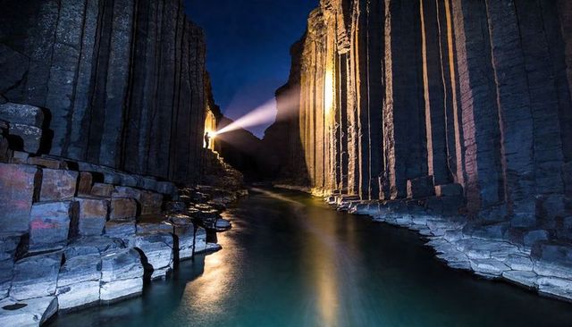 Dramatic hiker beaming flashlight across basalt column canyon at night river reflections