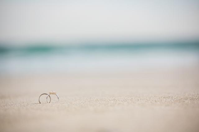 Wedding rings placed on sandy beach with ocean waves in background. Ideal for use in wedding invitations, engagement announcements, romantic getaway promotions, and beach-themed wedding decor.