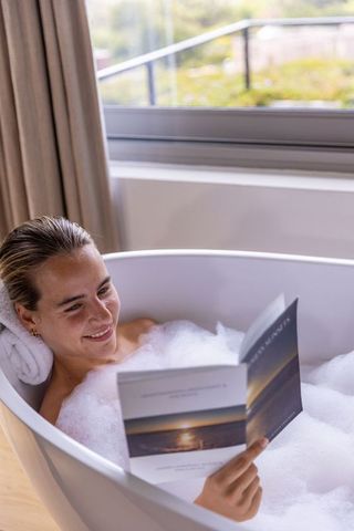 Woman reclining in freestanding bubble bath reading paperback, relaxing by sunlit window