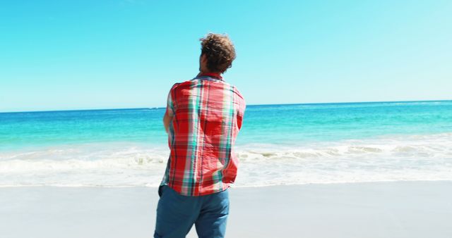 Man standing at the edge of the ocean on a sunny day, looking out at the blue water. Appearing relaxed and enjoying the tranquility of the beach. Ideal for travel, vacation, or leisure themes, and promoting outdoor activities and a laid-back lifestyle.