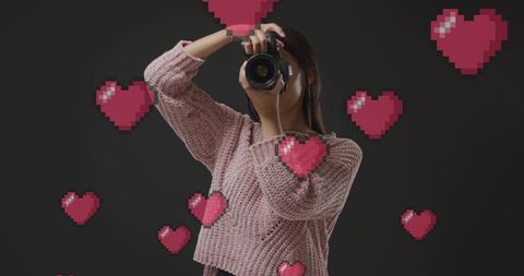 Woman photographer capturing shot with pixel heart overlay, pink chunky sweater portrait