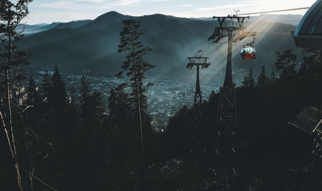 A cable car is ascending above a dense forest and mountainous area during sunrise. Rays of light penetrate through the mist, creating a serene and picturesque view. Perfect for promoting travel destinations, adventure tourism, nature retreats, and sightseeing excursions.