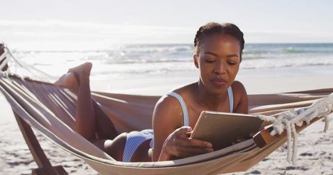 Woman relaxing in hammock on sunny beach reading tablet in light blue striped swimsuit