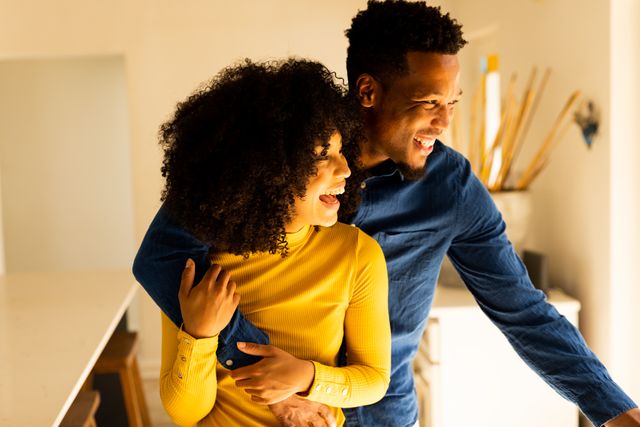 This image shows a happy biracial couple embracing and laughing at home. It can be used for promoting inclusivity, domestic life, leisure time, and togetherness. Ideal for advertisements, blogs, and social media posts focusing on relationships, love, and family life.