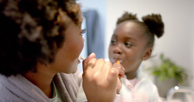 A woman and her young daughter are brushing their teeth together in a bathroom, creating a moment of bonding and teaching good health habits. This image highlights the themes of family interaction, daily hygiene, and parental care. Great for use in advertisements, parenting blogs, health and wellness articles, or dental care promotions.
