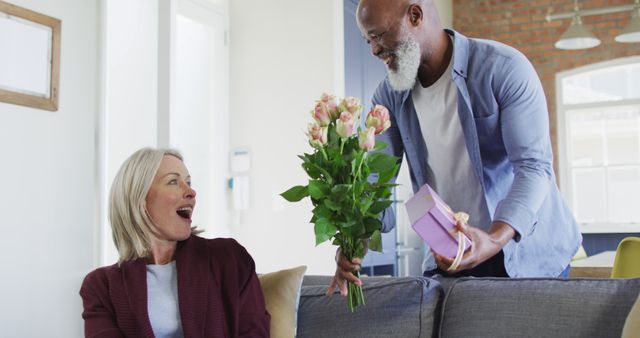Happy senior diverse couple in living room sitting on sofa, giving flowers and present. retirement lifestyle, spending time at home.