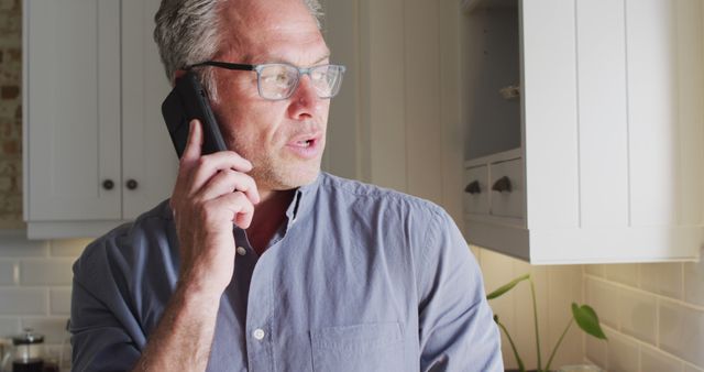 Mature man with glasses wearing casual shirt talking on phone in modern kitchen, looking concerned. Suitable for depicting business or personal communications, remote work, home environments, or lifestyle contexts.