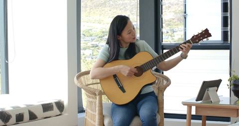 Asian woman playing acoustic guitar in sunlit modern living room with tablet