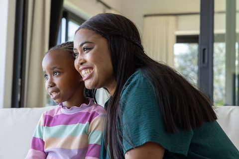 African american mother and daughter smiling together on sunlit cozy couch in living room