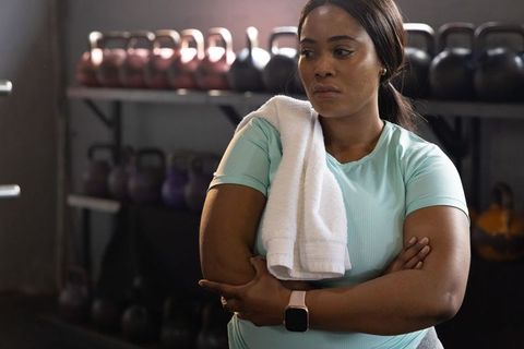 Focused african american woman standing in gym holding towel by kettlebells and smartwatch