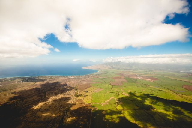 Aerial view of Hawaiian coastline displaying lush green fields merging with pristine ocean waters under a dramatic sky with scattered clouds. Ideal for travel brochures, tourism websites, or advertisements promoting tropical destinations and nature explorations.