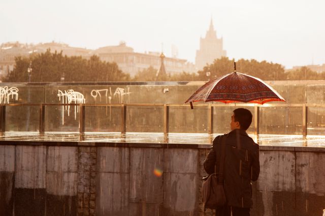 A man with a red umbrella is walking in the rain during sunset in an urban area with a city skyline in the background. Ideal for use in articles or advertisements about urban living, seasonal weather, solitude, and evening activities.
