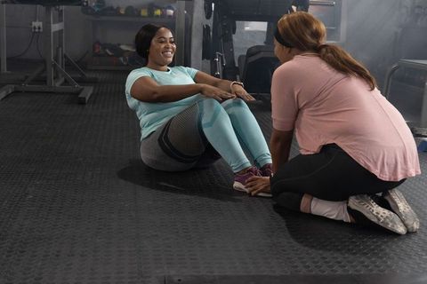 Two women doing partner sit-ups in gym holding feet, smiling during core workout