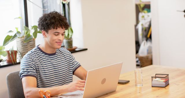 Young man with curly hair working on laptop in modern home office. Surrounded by plants and natural light, with organized desk. Perfect for themes of remote work, productivity, modern living, or technology. Ideal for articles, blogs, and advertisements related to work-from-home setups, home office design, or digital lifestyle.