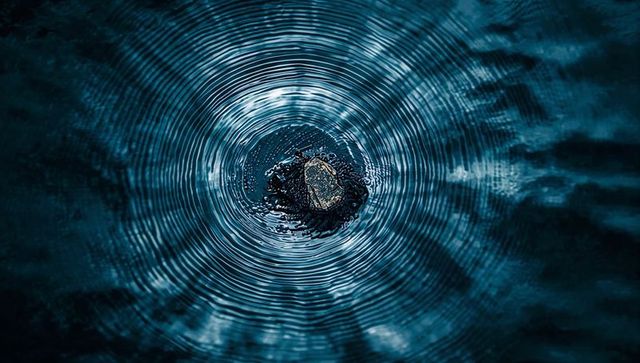 Dark pebble creating concentric ripples on teal water with radial symmetry and stillness