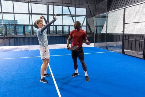 Two male athletes playing padel on blue indoor court inside modern glass-walled facility