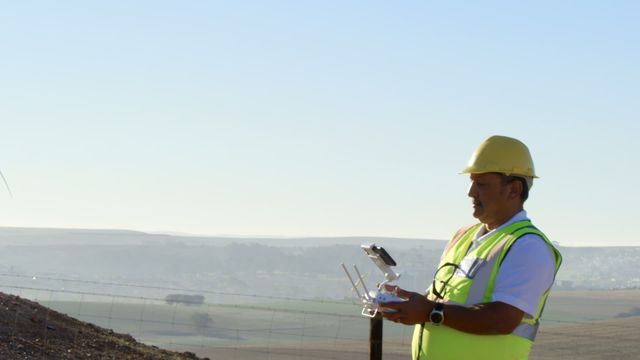 A male engineer in a high visibility vest and hard hat is operating a drone near a wind turbine. The landscape offers a clear horizon, symbolizing clean and sustainable energy initiatives. Ideal for use in advertising renewable energy projects, showcasing modern engineering practices, and highlighting technological advancements in environmental studies.