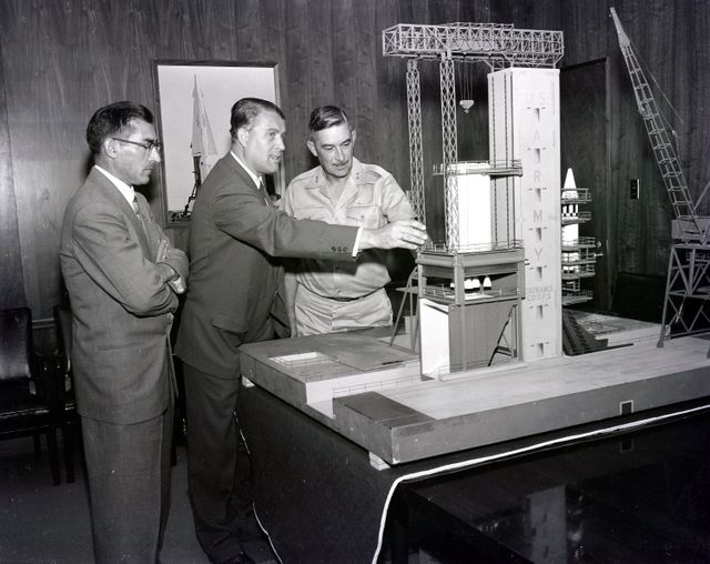 Image shows historically significant meeting discussing the S-1B Test Stand model during the 1950s. Key figures involved in the U.S. rocket program including Karl L. Heimburg, Dr. Wernher von Braun, and Major General John B. Medaris are visible. Great for historical articles, documentaries on missile development, space race education materials, and features on space exploration history.