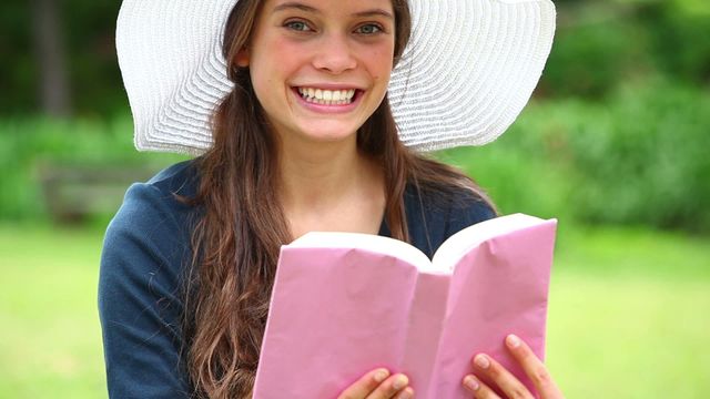 A young woman wearing a wide-brimmed hat is sitting in a park with a big smile, holding a book. Perfect for use in advertising for lifestyle, relaxation, or outdoor activities. Suitable for articles about leisure reading, enjoying nature, or promoting cheerful moments outdoors.