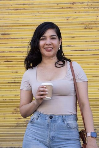 Smiling woman holding coffee by yellow roll-up door, urban casual fashion portrait