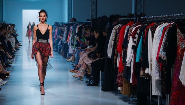 Runway model walking in black bodysuit and printed high-slit skirt with racks and audience