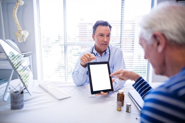 Doctor and senior patient are using a digital tablet during a consultation in a clinic. The doctor is explaining something on the tablet while the patient listens attentively. There are medication bottles on the table, indicating a discussion about prescriptions or treatment plans. This image can be used for healthcare websites, medical blogs, articles on elderly care, or promotional materials for medical services.
