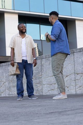 Diverse male coworkers chatting on urban office plaza holding lunch bag and reusable cup