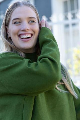 Smiling young woman in green sweater tucking hair – candid urban headshot, daylight