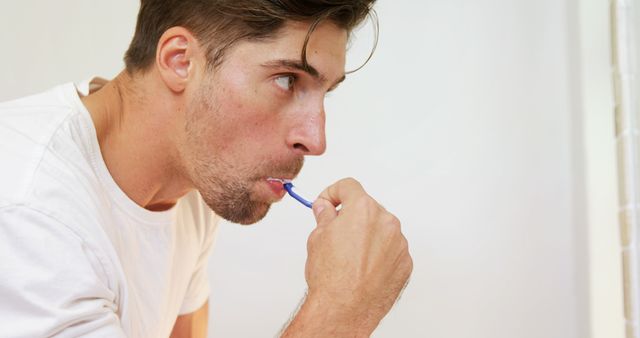 Young man is brushing teeth in a bright bathroom, contributing to daily dental hygiene and oral care. Ideal for themes related to health, morning routines, grooming, dental care products, and cleanliness.