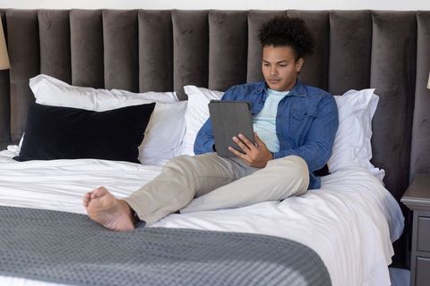 Mid-adult man relaxing on bed browsing tablet in modern cozy bedroom with tufted headboard