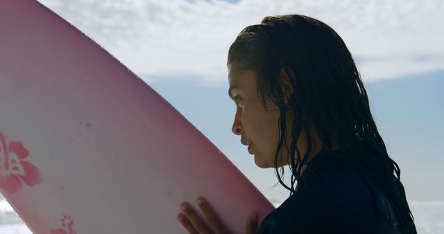 Young female surfer holding pink surfboard, looking pensive at beach, sea in background. Useful for themes on surfing lifestyle, outdoor adventures, beach activities, summer sports, and female empowerment in sports.