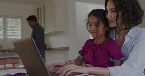 Mother guiding daughter on laptop at kitchen table while partner works in background