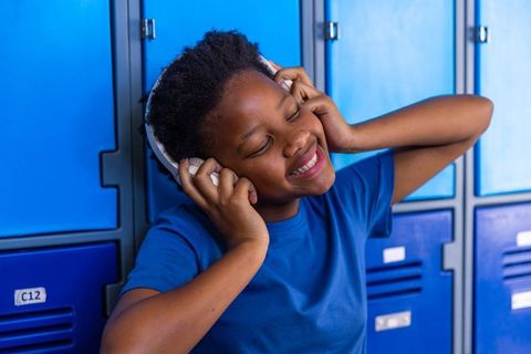 African american teen listening to music on headphones while leaning on blue lockers
