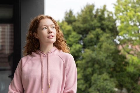 Redhead woman wearing pink hoodie looking upward on balcony with soft green bokeh